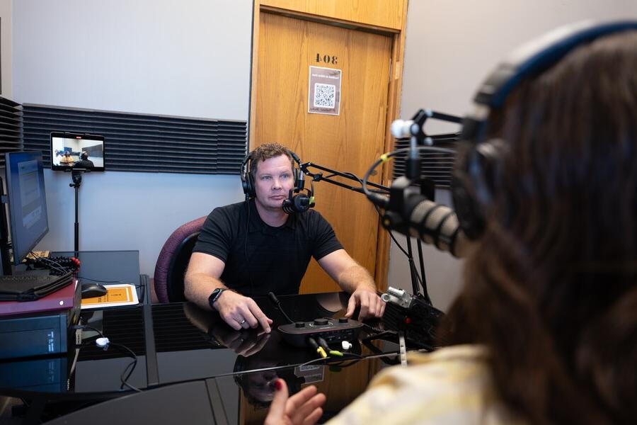 A man sits in a small room with headphones on in front of a microphone and other podcast equipment.